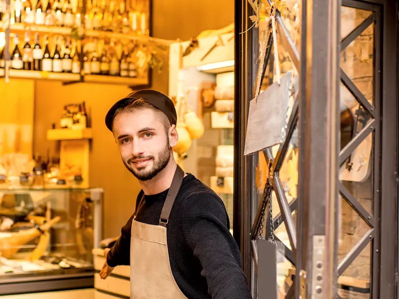A shopkeeper in an apron welcomes people in at the door of his shop.
